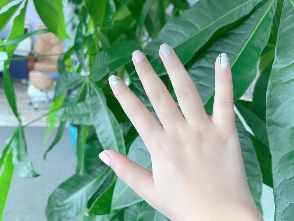 A hand with short nails in sheer nude and milky white, one finished with a thin black cross-hatch and one with a single stone