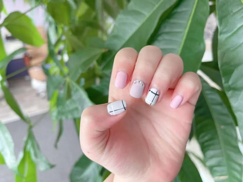 Four fingernails close up — baby pink, a milky nude set with three small stones, and white with fine black lines drawn across it